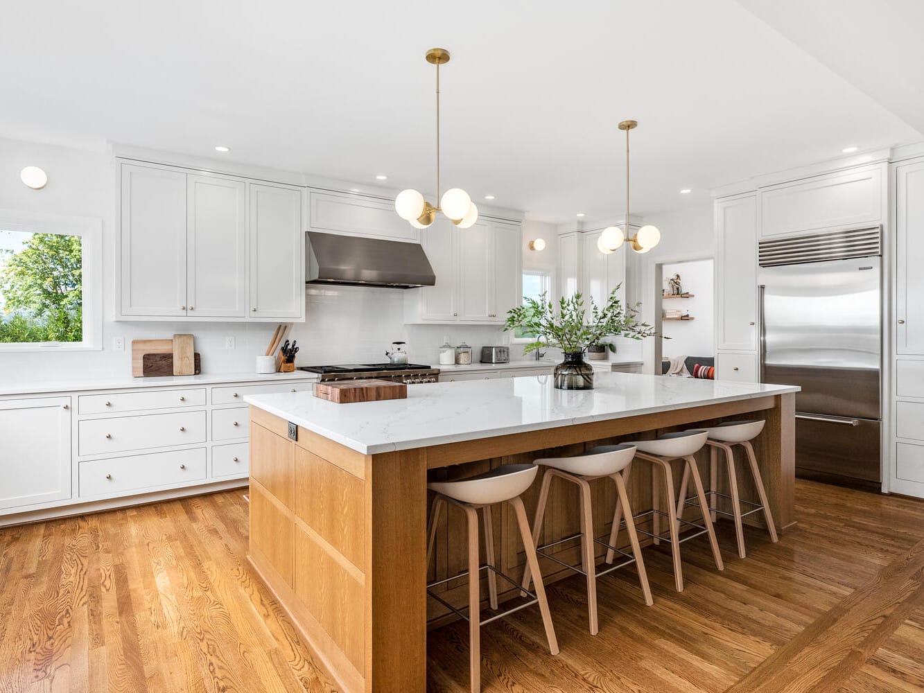 Modern kitchen with white cabinets and a large wooden island, reminiscent of Portland, Oregon's sleek design style. Three white bar stools line the island, topped with a vase of greenery. Stainless steel appliances and pendant lights complete the bright space on hardwood flooring.