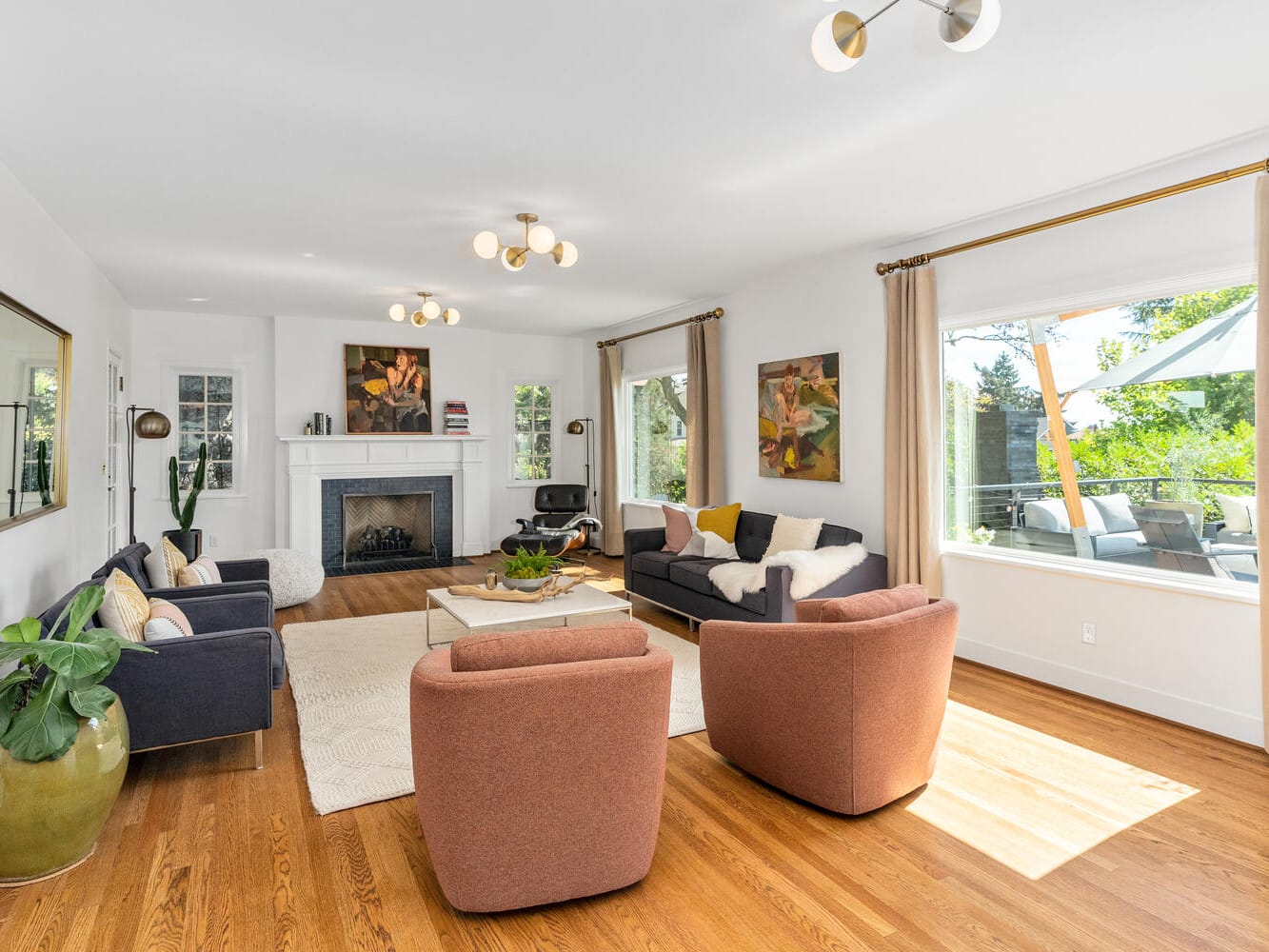 A spacious living room in Portland, Oregon, features large windows, two gray sofas, two pink armchairs, and a coffee table. The fireplace showcases artwork above it, while plants and neutral-colored decor enhance the space. Natural light floods the room.
