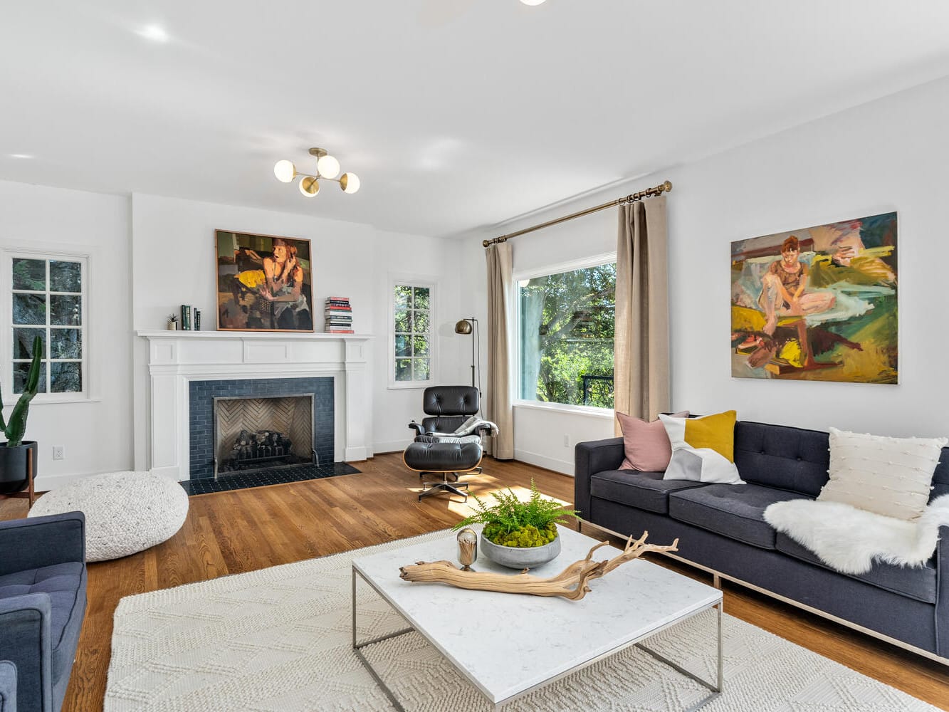 A modern living room in Portland, Oregon, features a fireplace, abstract art, and an Eames chair. The space boasts dark gray sofas with colorful cushions, a large window showcasing tree views, a white rug, and a marble coffee table adorned with decorative driftwood and a plant.