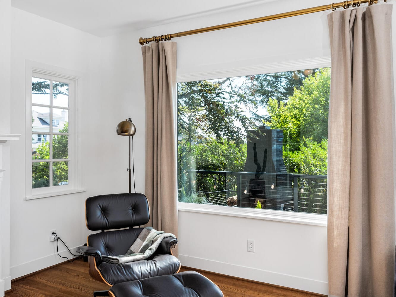 A cozy Portland, Oregon living room features a black leather lounge chair and ottoman near a large window. Beige curtains frame the view of lush greenery outside. A brass floor lamp stands nearby on the hardwood floor, while a stack of books rests on the mantel.