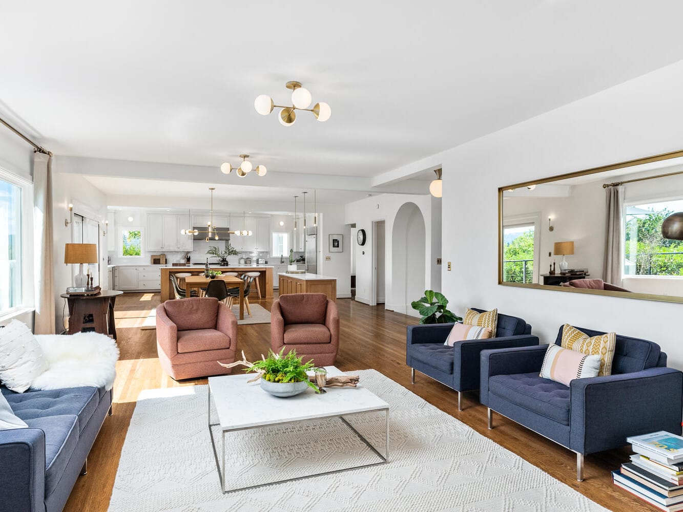 Spacious living room in Portland, Oregon with modern decor featuring a large mirror, blue and pink armchairs, a marble coffee table, and a plush rug. An open kitchen with wooden dining furniture is visible in the background, illuminated by stylish pendant lights.