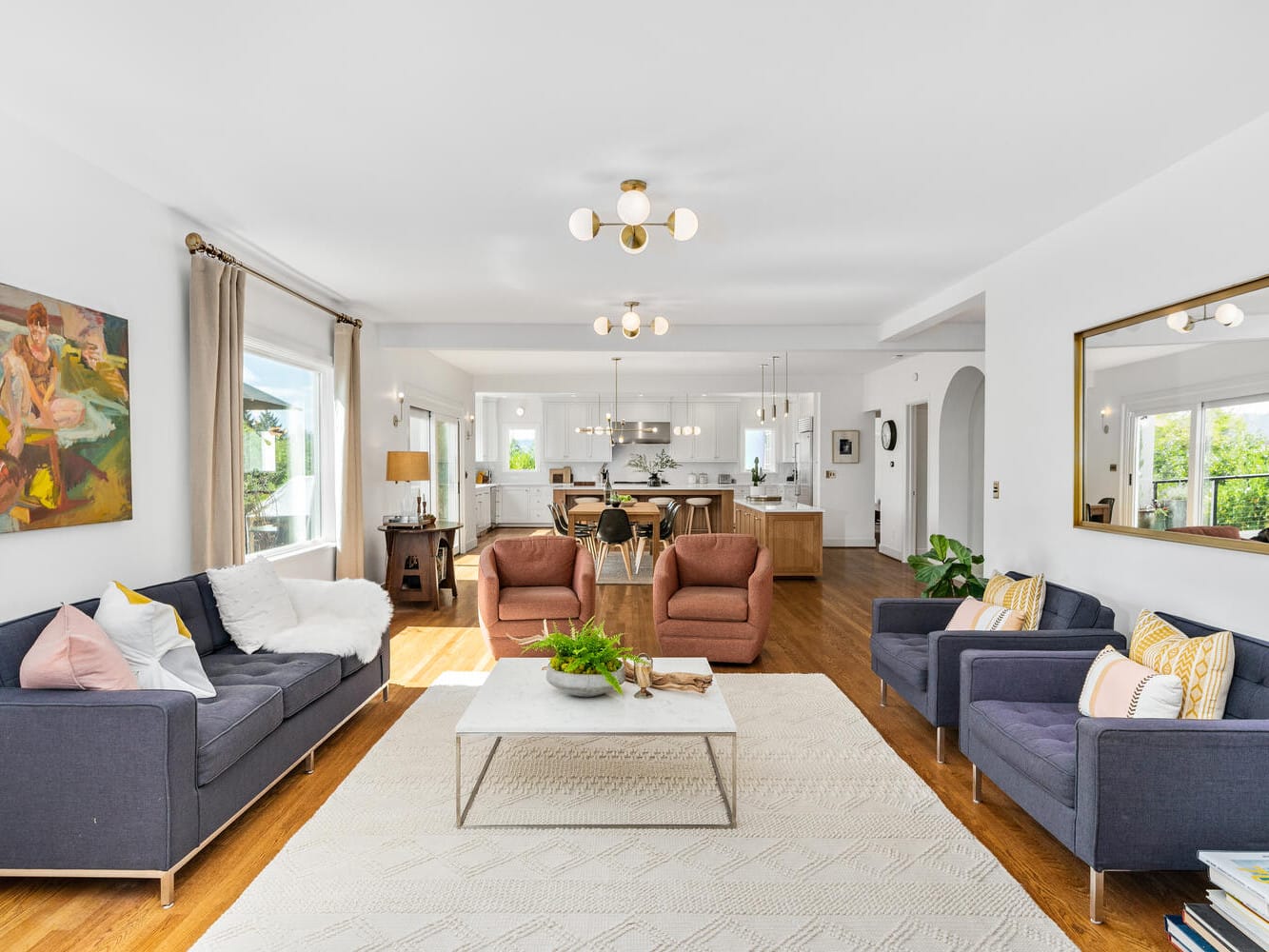 A bright living room in Portland, Oregon, boasts modern decor with two dark sofas, pink chairs, and a coffee table adorned with plants. A large mirror enhances the space that seamlessly opens to a dining area and kitchen featuring white walls and wooden floors.