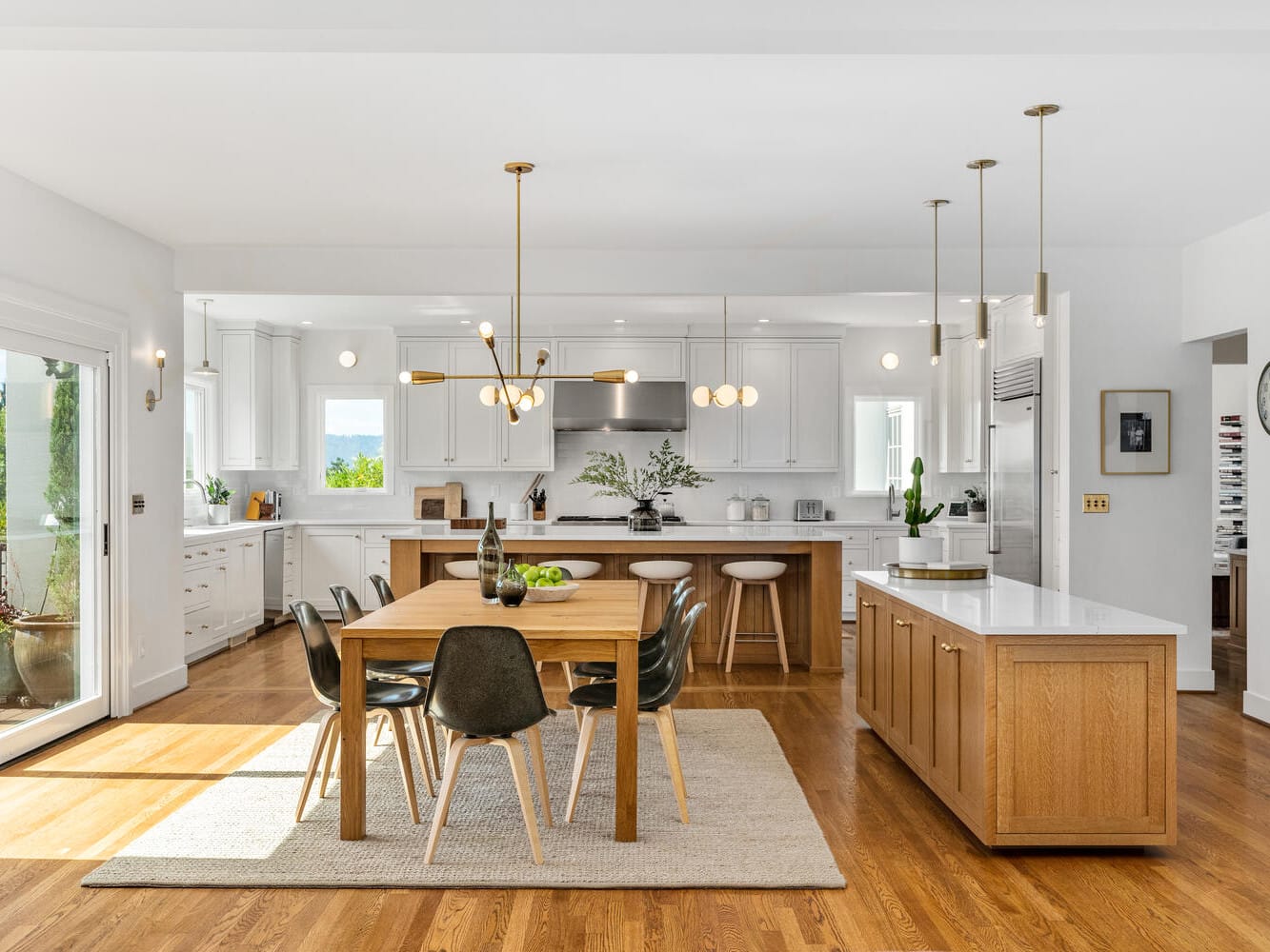 A spacious, modern kitchen in Portland, Oregon, with hardwood floors and white cabinets. It features a wooden dining table with black chairs, an island with bar stools, pendant lighting, and large windows letting in natural light. White walls and greenery add to the decor.