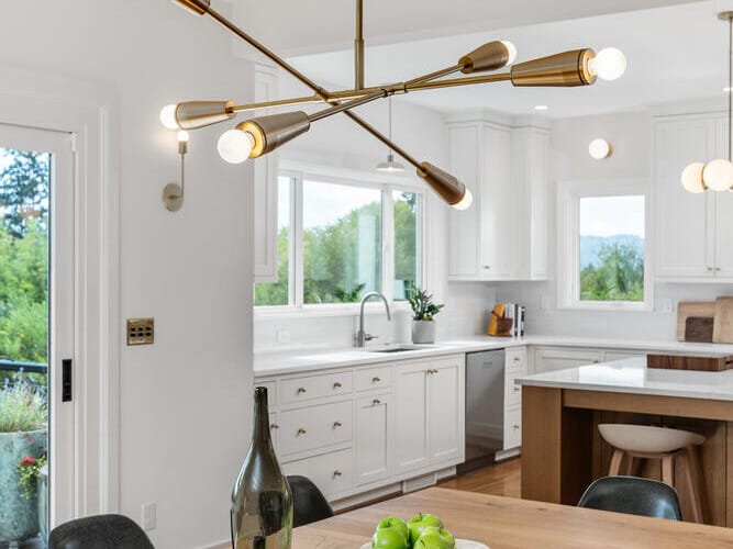 A modern kitchen in Portland, Oregon, with a wooden dining table featuring a fruit bowl filled with green apples and a decorative bottle. A contemporary chandelier hangs above. The room has white cabinets, large windows, and a view of lush greenery outside.