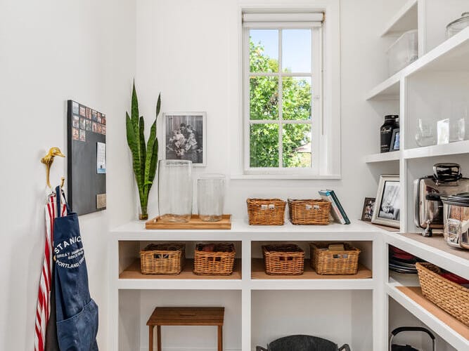 A tidy Portland pantry with white shelves hosts wicker baskets, a dark apron on a wall hook, and various kitchen items. A small window with a blind reveals lush greenery outside. The hardwood floor is immaculate, and a snake plant from Oregon sits gracefully on the counter.