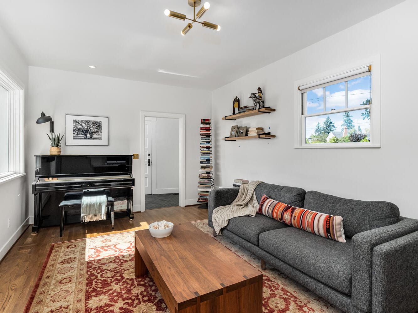 A cozy living room in Portland, Oregon features a gray sofa with colorful cushions, a wooden coffee table, and a piano in the corner. Natural light streams through the windows onto shelves filled with books and decor. A red patterned rug adds warmth to the inviting space.