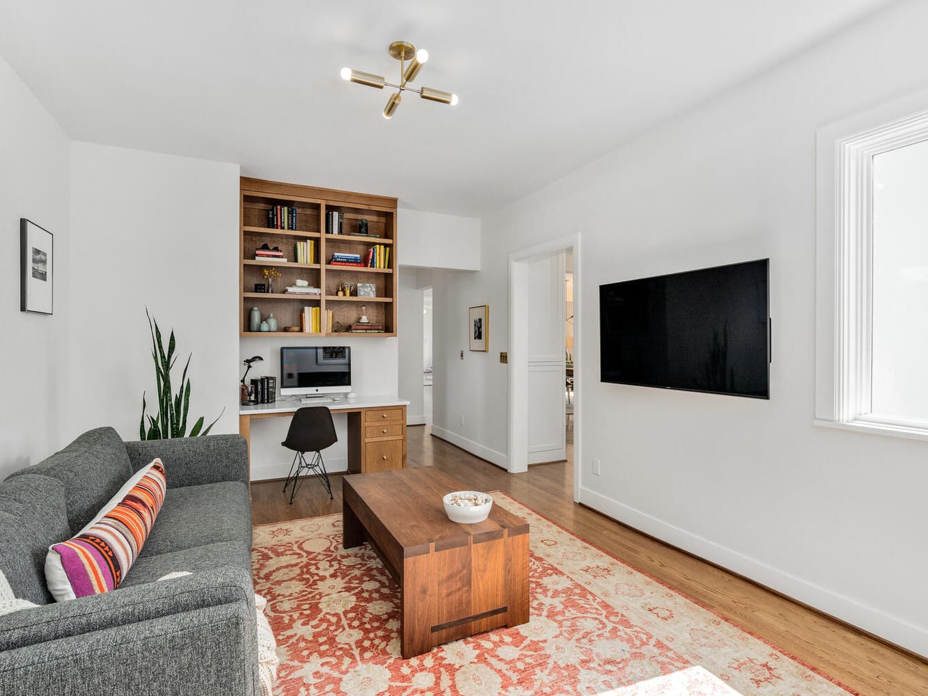 A modern living room in Portland, Oregon features a gray sofa with colorful pillows, a wooden coffee table, and a wall-mounted TV. A built-in wooden shelf with books and desk occupies the corner. A plant and textured rug add warmth to this bright Pacific Northwest space.