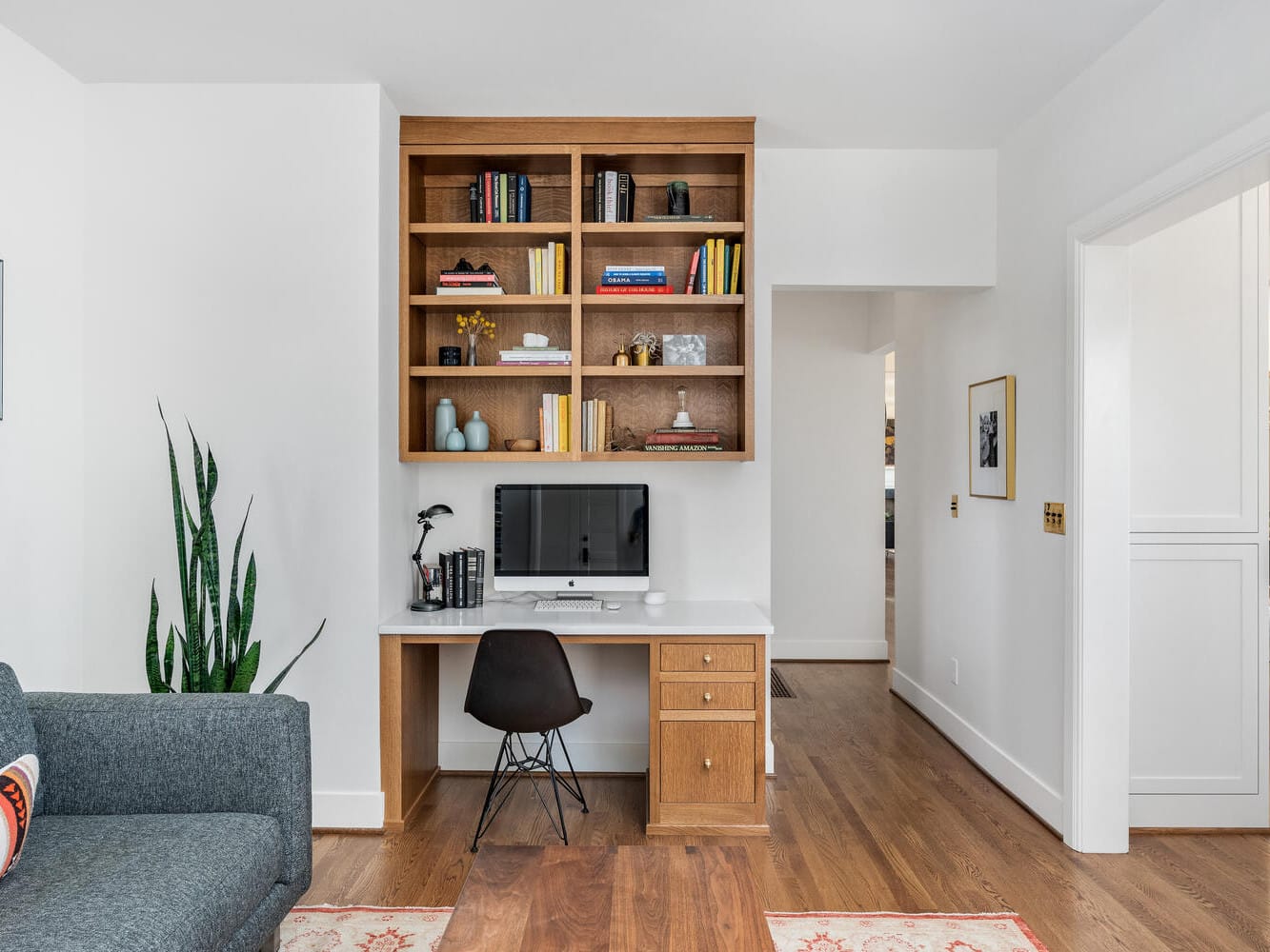 A modern Portland, Oregon home office with a wooden desk and built-in shelves. The computer sits on the desk, surrounded by books and decorations. A black chair is positioned at the desk. A large potted plant and a gray sofa are nearby, with framed photos on the walls.