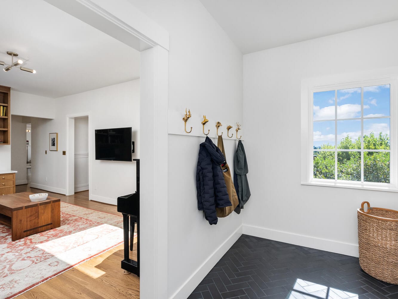 A bright and modern foyer in Portland, Oregon, features white walls and a window revealing a clear blue sky, with a coat rack holding jackets and a woven basket. The adjoining room showcases a wooden coffee table and wall-mounted TV.