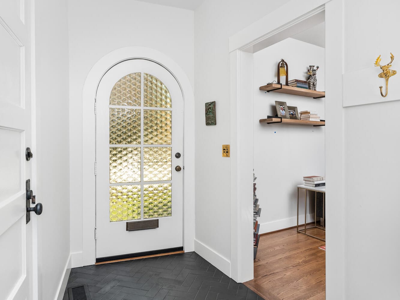 A bright entryway in Portland, Oregon, showcases a white arched door with decorative glass panels. The room features white walls, a dark herringbone floor, and offers a glimpse of an adjacent office filled with shelves, books, and a desk. A decorative wall hook adds charm to the space.