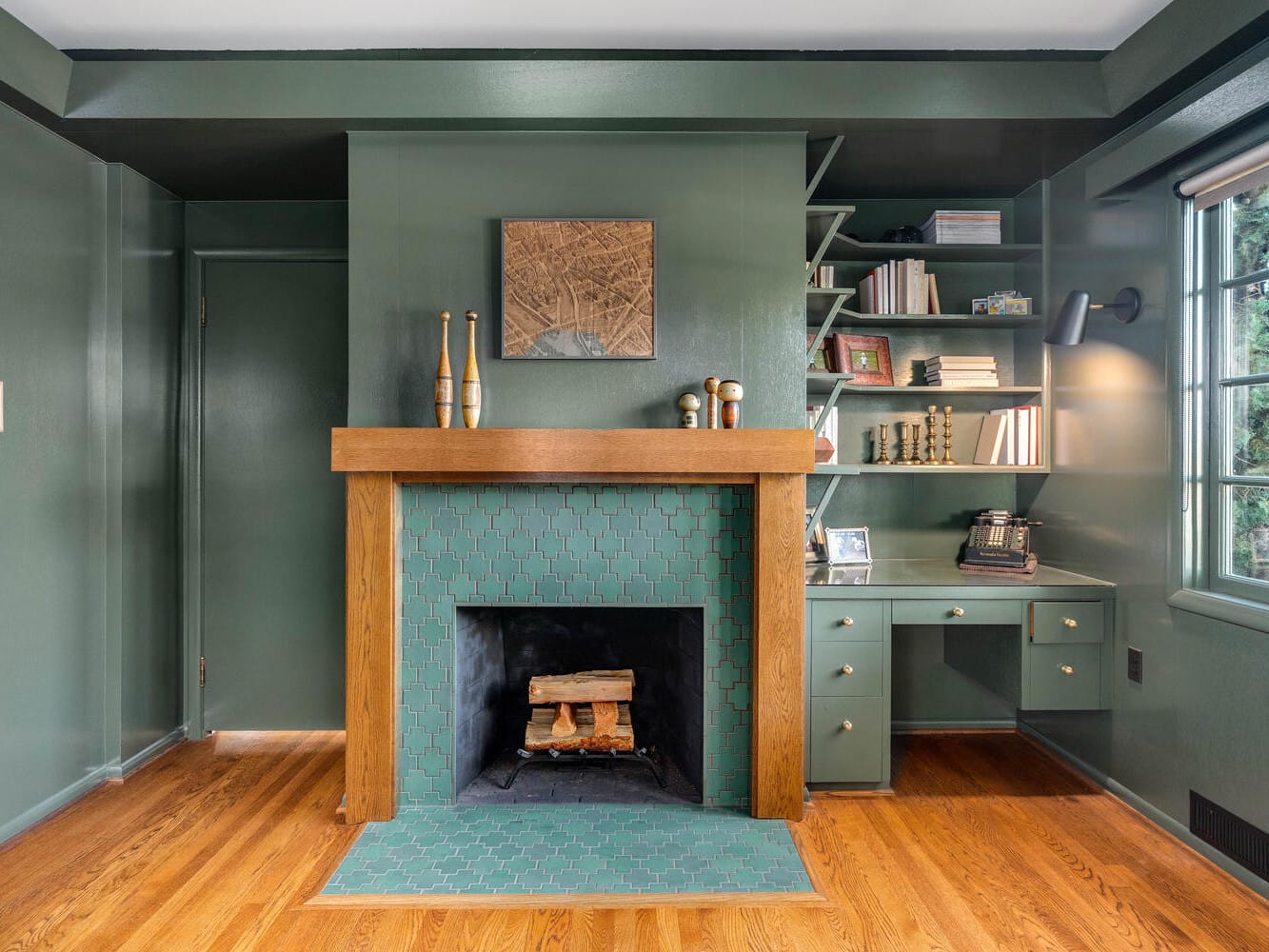 A cozy Portland room showcases a green, tiled fireplace with a wooden mantel. Shelves adorned with books and decor enhance its charm. To the right, a built-in desk features a vintage phone. The green-painted walls and ceiling beautifully complement the wooden floor and natural light from the window.