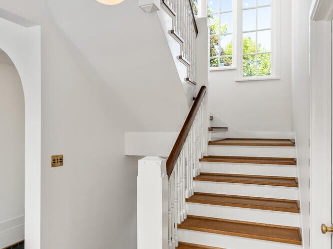 A bright staircase with white walls and wooden steps leads upward in a Portland, Oregon home. A large window fills the space with natural light, while a modern round light fixture hangs from the ceiling. An arched doorway is visible on the left.