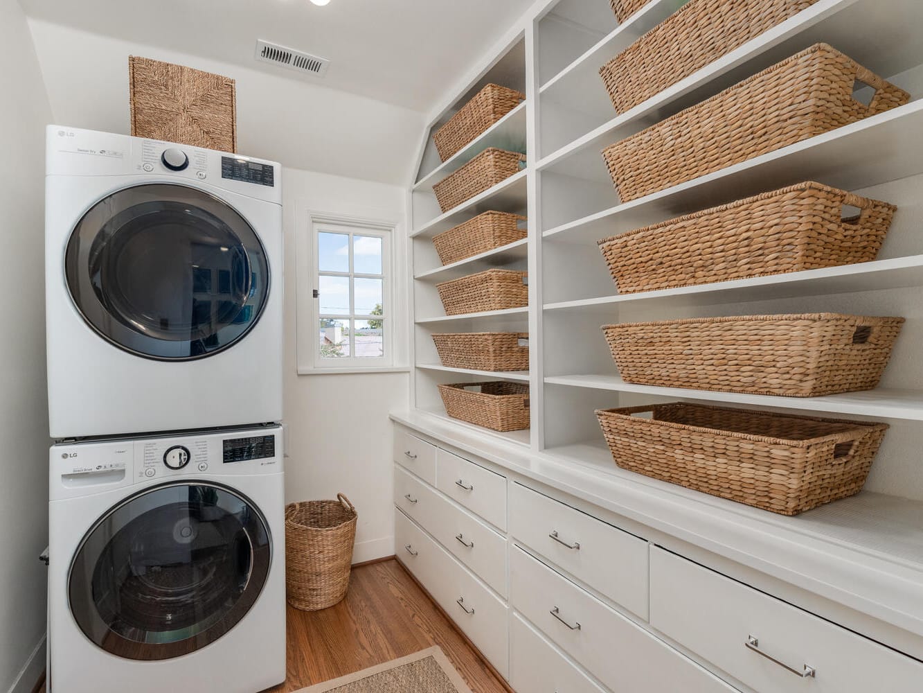 A cozy Portland, Oregon laundry room features a stacked washer and dryer on the left. On the right, white shelves are filled with woven baskets, all bathed in natural light from a small window in the background. The warm wood floors complete this charming space.