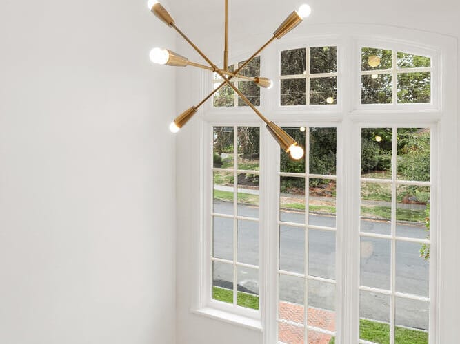 A modern staircase in Portland, Oregon features a wooden railing, white walls, and a large window with a street view. A gold chandelier with exposed bulbs hangs from the ceiling, adding a stylish touch to the bright space.