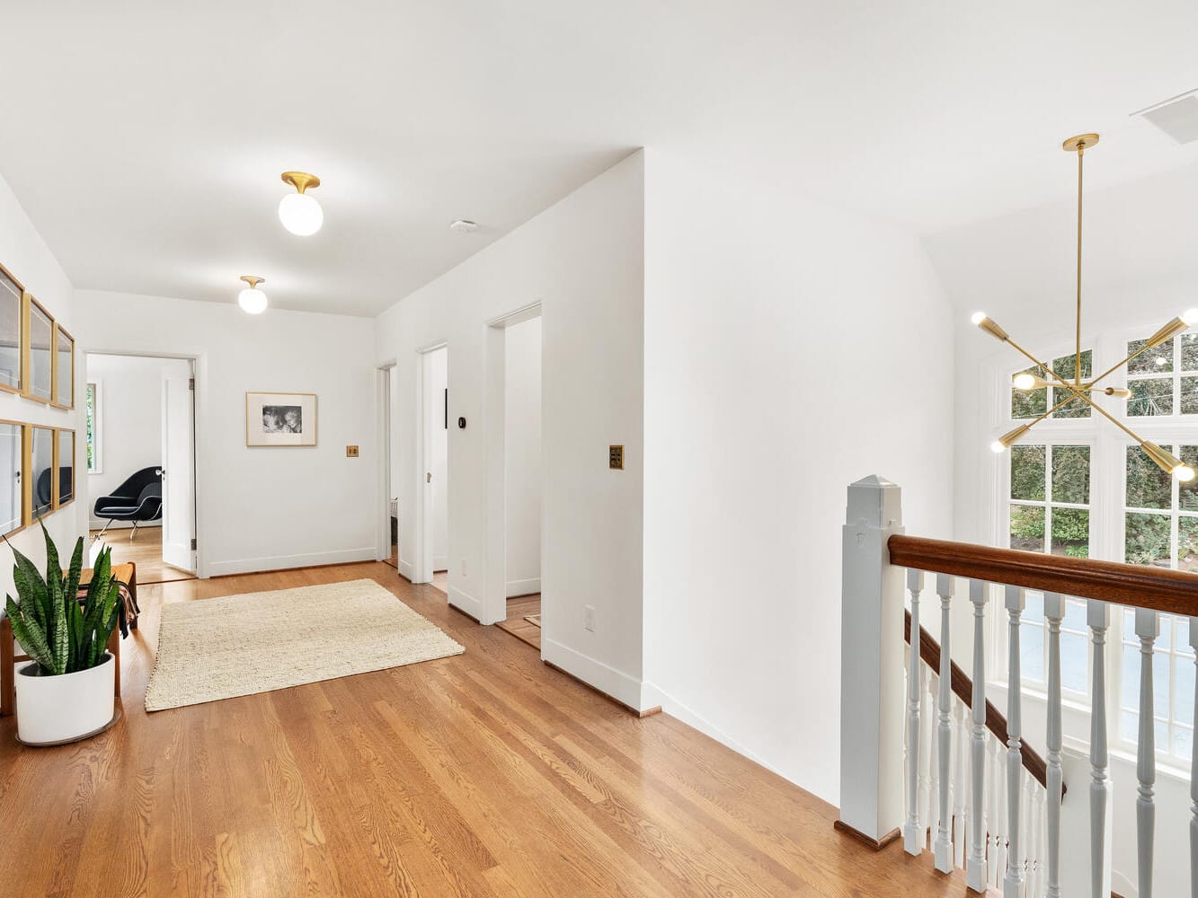 A bright, modern hallway in a Portland, Oregon home showcases wooden floors, white walls, and multiple round ceiling lights. It features framed art on the left wall, a potted plant on the floor, and offers a view of a staircase with a large window.