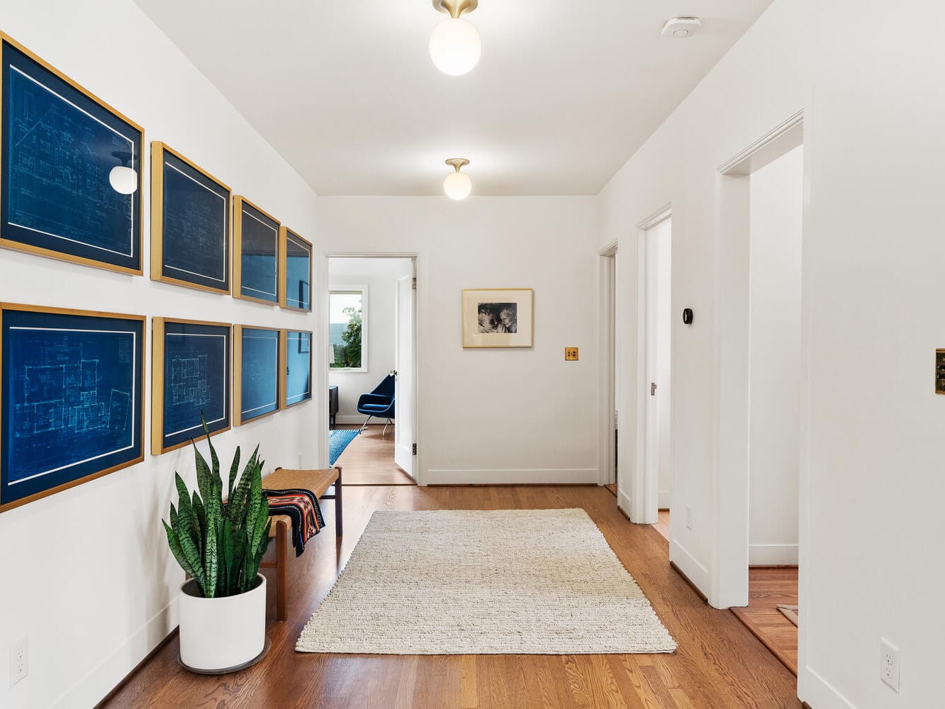 A bright hallway with wooden floors, reminiscent of classic Portland, Oregon style, is decorated with framed blueprints on the left wall. A large rug covers part of the floor. A potted plant and small bench are on the left, leading to a doorway at the end of the hall.