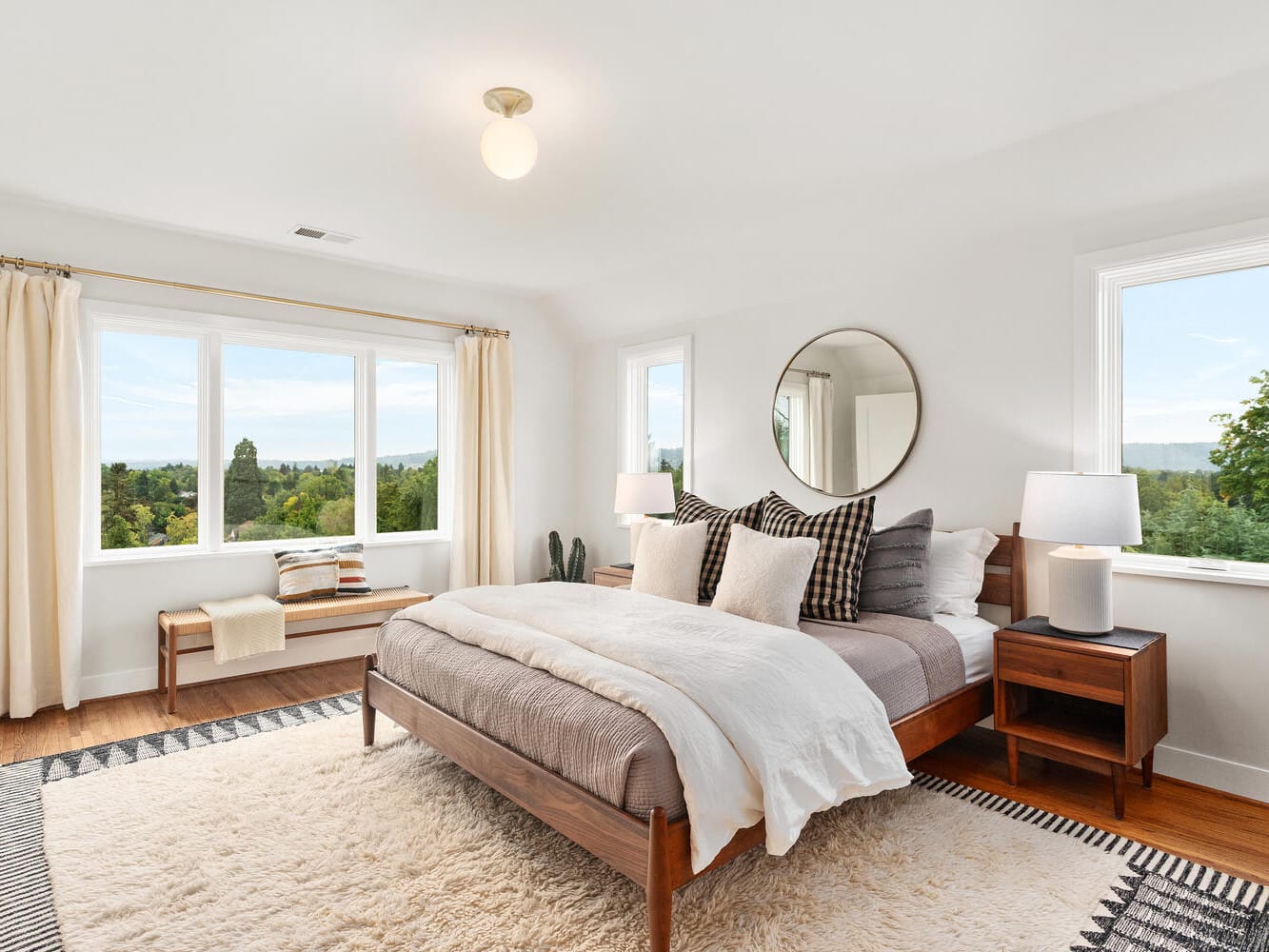 A bright and airy bedroom in Portland, Oregon, with a large window showcasing a scenic view. The room features a bed with layered pillows and a neutral-toned duvet, complemented by a round mirror above the wooden nightstand, lamps, a rug, and a bench under the window.