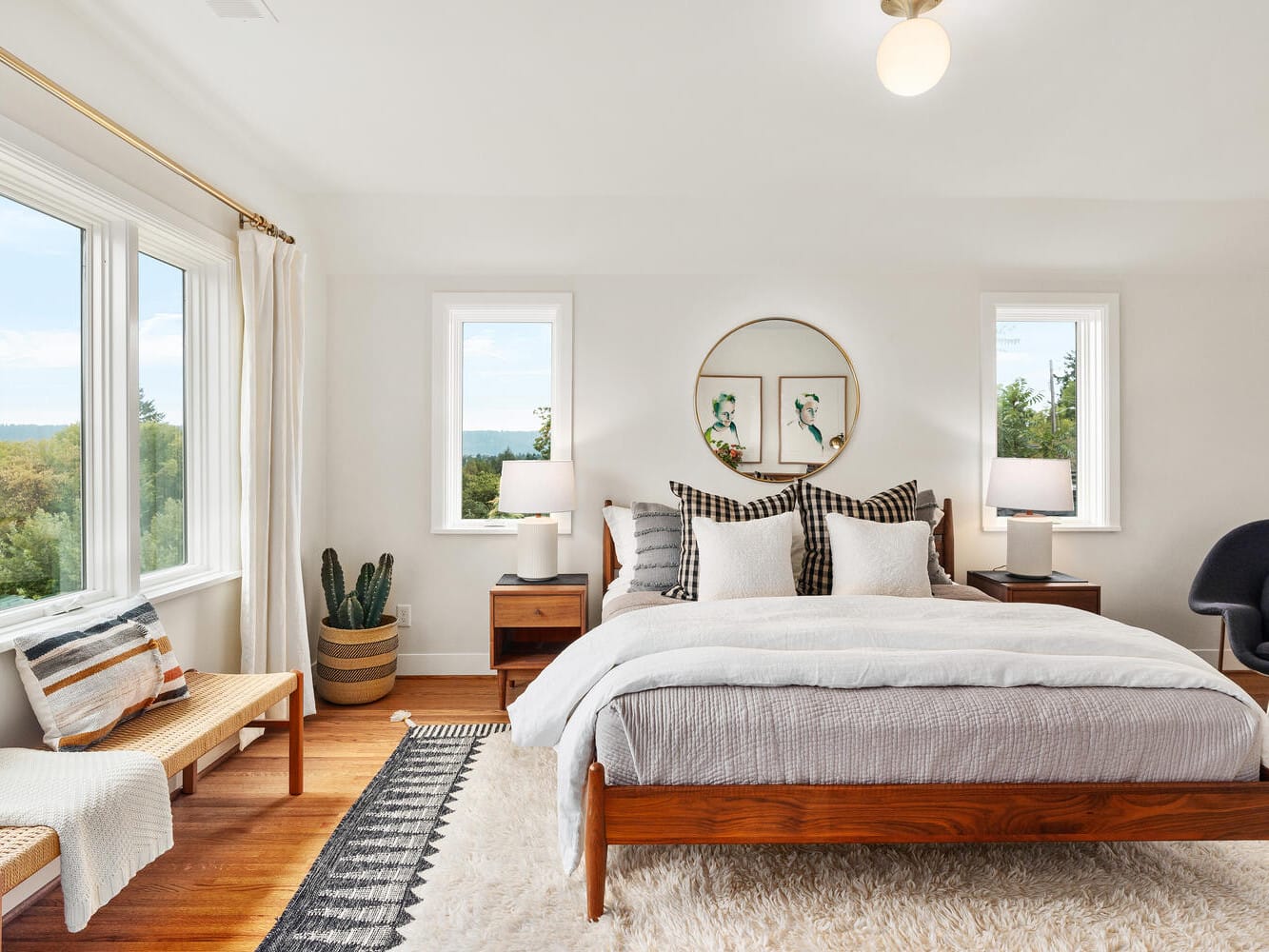 A bright bedroom in Portland, Oregon, boasts a large window and a wooden bed with white and plaid bedding. Flanked by two bedside tables with lamps, it features a round mirror and potted plant for decor. A plush rug graces the hardwood floor, while a bench sits by the window.