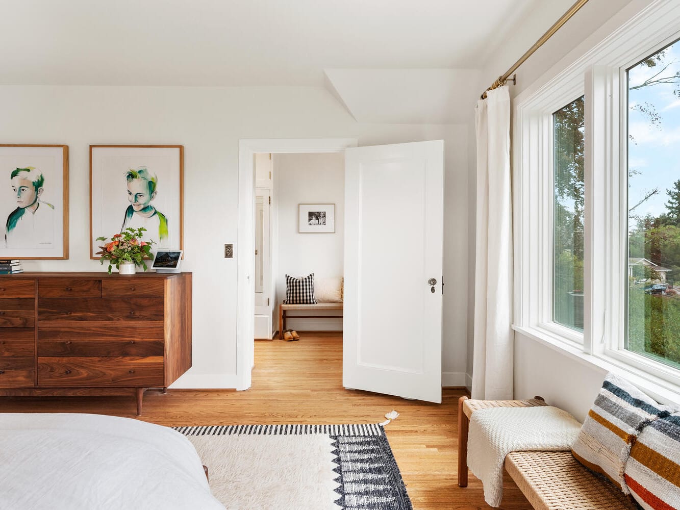 A bright bedroom in Portland, Oregon features a large window, a wooden dresser adorned with decorative items, and two framed artworks above it. A partially open door leads to another room. A woven bench with cushions sits atop a patterned rug that complements the warm wooden floor and light walls.