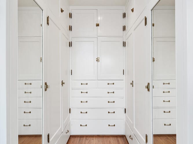 A walk-in closet in Portland, Oregon, showcases white cabinets and drawers on wooden floors. Full-length mirrors line the walls, and brass handles adorn every drawer and cabinet. Recessed lighting brightens the elegant space.