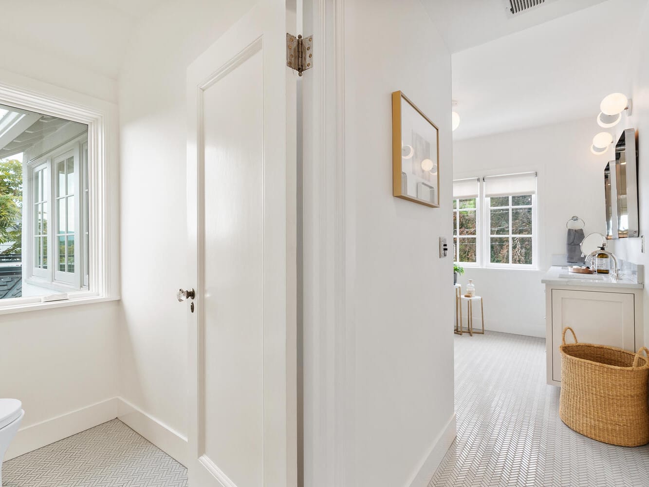 Bright, modern bathroom in Portland, Oregon, with a window, white walls, and herringbone-patterned floor. Features a vanity with a mirror, a wicker laundry basket, and soft lighting. The space is divided by a partial wall with a closed door.