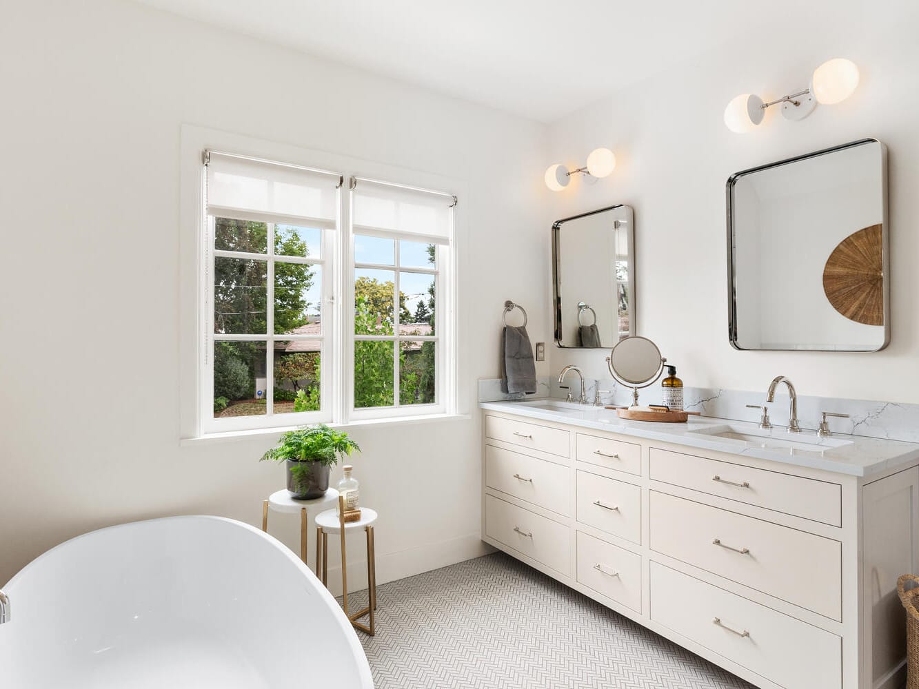 Bright, airy bathroom in a Portland, Oregon style, featuring a large white bathtub, double vanity with mirrors, and modern light fixtures. A window offers a view of lush greenery outside. A small plant graces a round side table, with a woven basket beside the vanity.