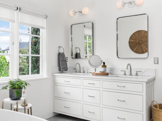 In a bright Portland, Oregon bathroom, a white double sink vanity with a marble countertop and mirrors adds elegance. A bathtub with a towel sits in the foreground as natural light streams through large windows. Minimalist decor is complemented by a woven basket beside the vanity.