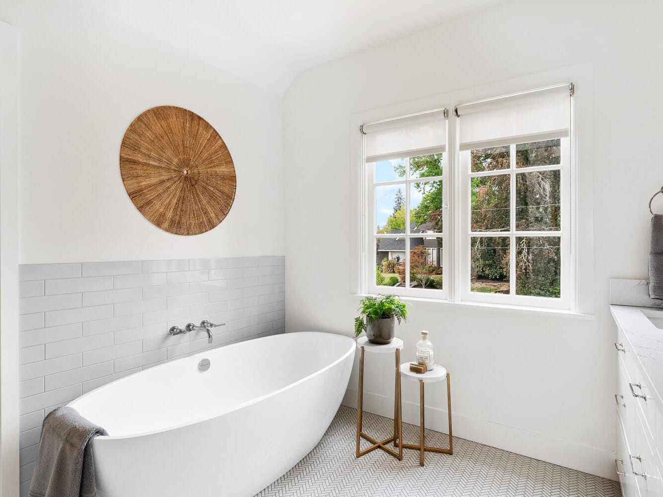 A bright, modern bathroom in Portland, Oregon features a freestanding white bathtub with a gray towel draped over the edge. A round, textured wall decoration is above the tub. Two small tables with a plant and soap sit by the window, overlooking a lush garden.