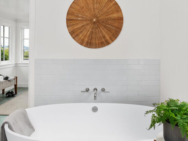 A minimalist bathroom in Portland, Oregon features a white freestanding bathtub with a gray towel draped over the edge. A round wicker decoration hangs on the wall above white tiles, while a potted plant sits gracefully on a small side table beside the tub.