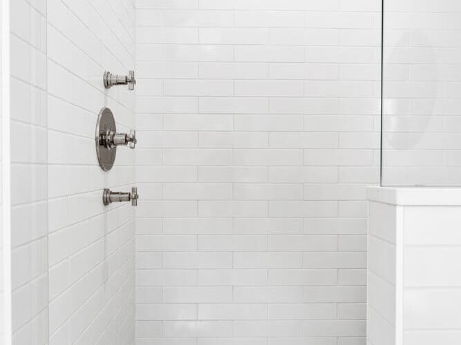 A modern shower in Portland, Oregon showcases white subway tiles and a chrome rain showerhead. The controls are mounted on the left wall, while the floor features a grey and white herringbone pattern. A glass partition is visible on the right, adding to its sleek design.