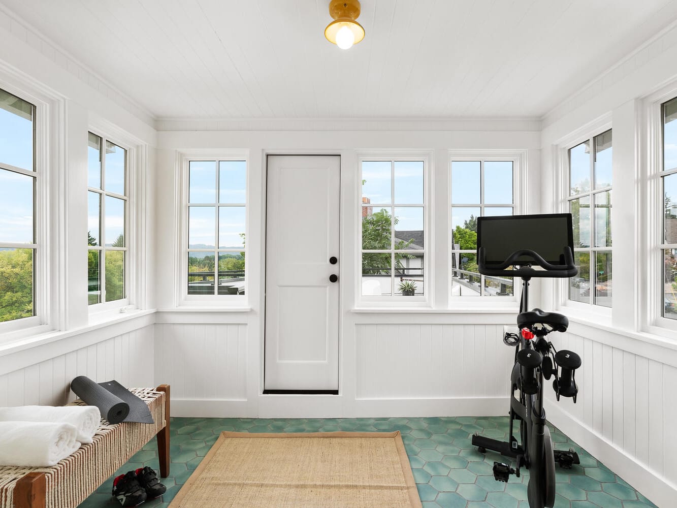 A bright exercise room in Portland, Oregon, features large windows and a stationary bike facing a wall-mounted screen. Nearby, a bench waits with towels and a yoga mat. Green hexagonal tiles cover the floor, and a central door leads outside.