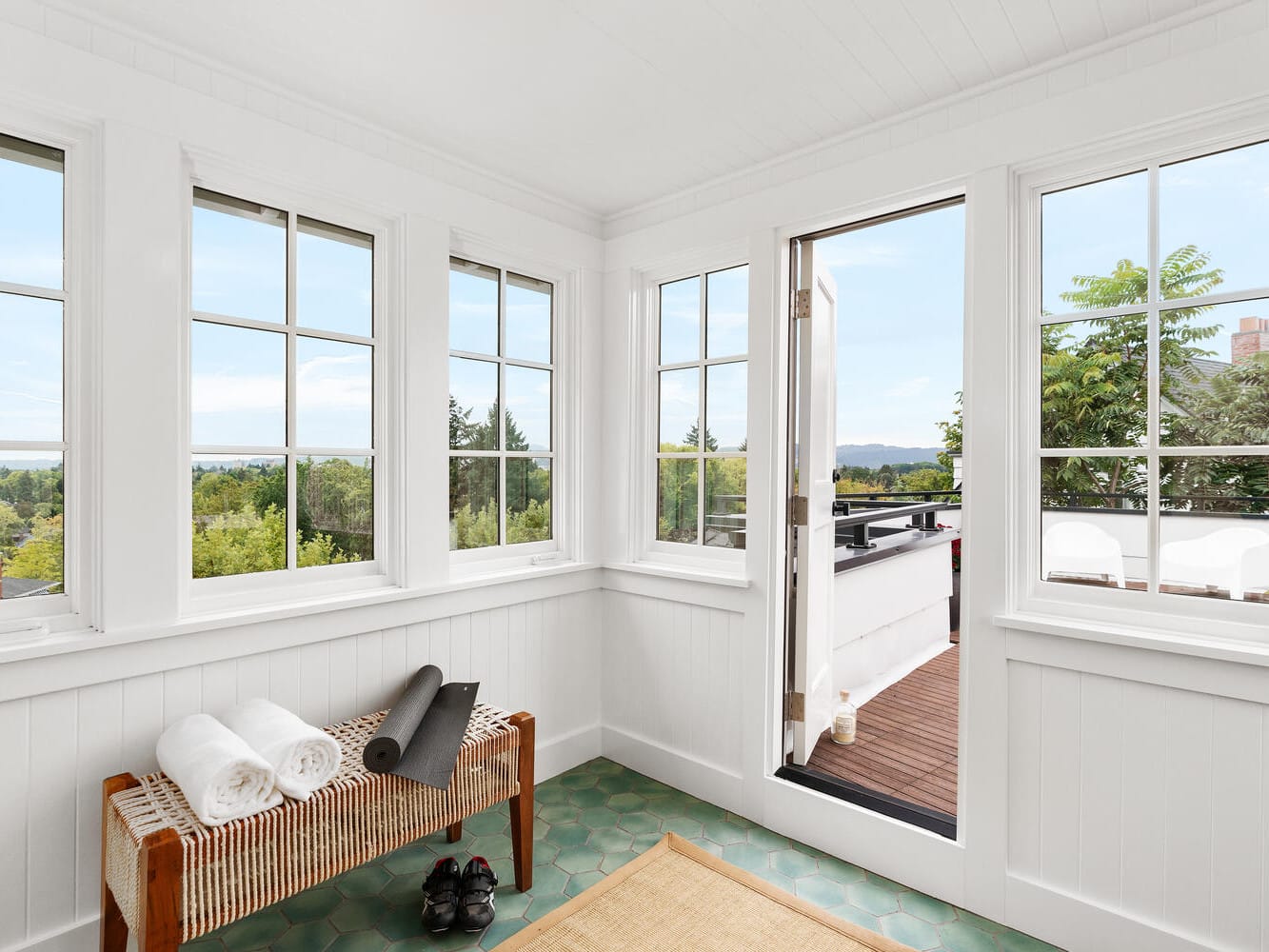 A bright sunroom with white walls and large windows offering a view of green trees and distant hills reminiscent of Portland, Oregon. The room features a wicker bench with rolled towels and a yoga mat, a pair of black shoes, and a door leading to a balcony with outdoor seating.