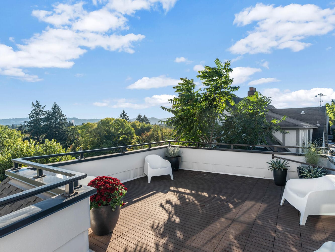 A rooftop terrace in Portland, Oregon, with wooden flooring features two white chairs and several potted plants, including a red flowering plant. The area is surrounded by a black railing and offers views of trees, hills, and a partly cloudy blue sky.