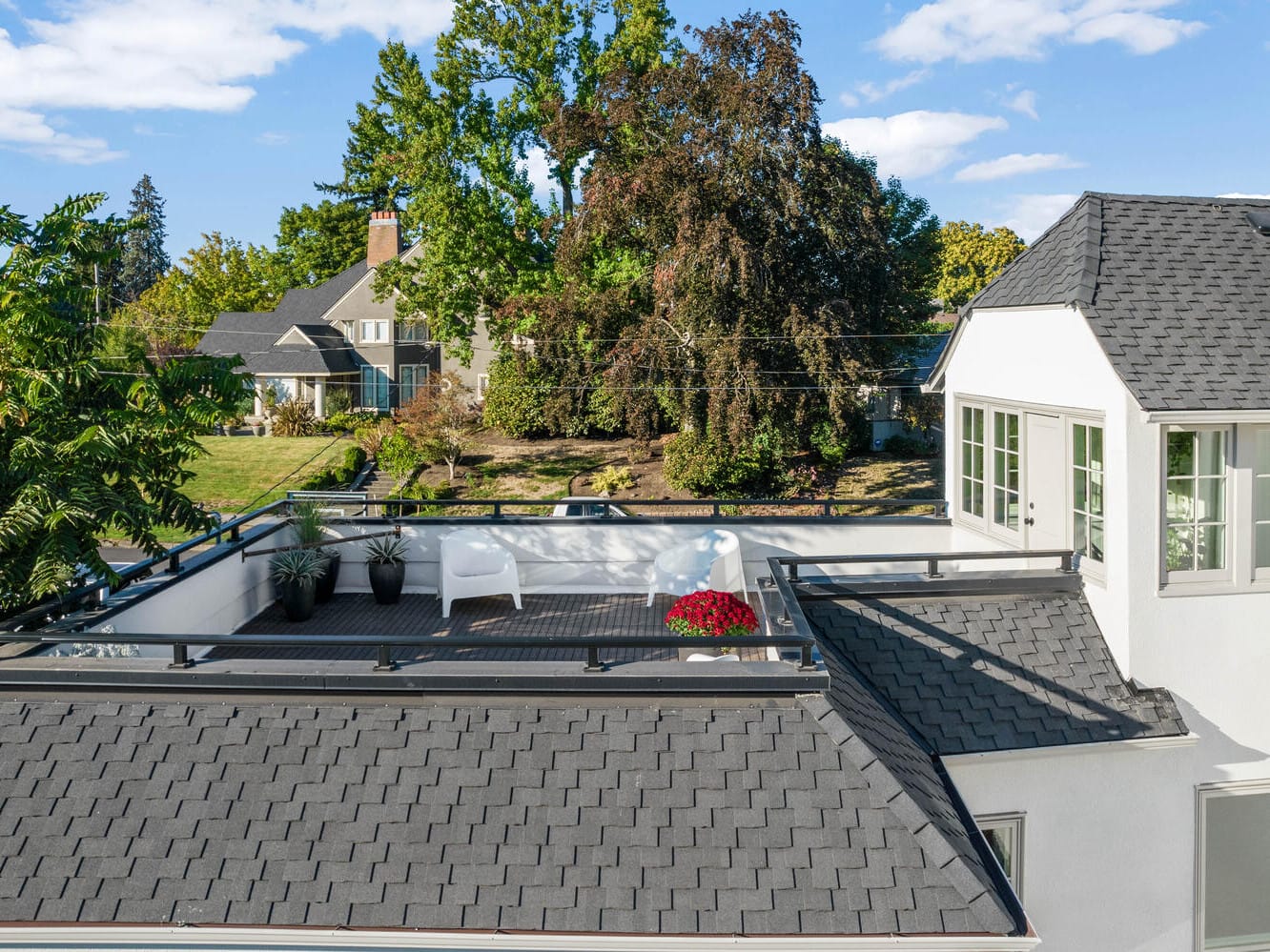 Aerial view of a white house in Portland, Oregon, featuring a black shingled roof and a rooftop deck. The deck hosts a white sofa, small table, potted plants, and a red flower pot. Trees and neighboring houses are visible under the blue sky with scattered clouds.