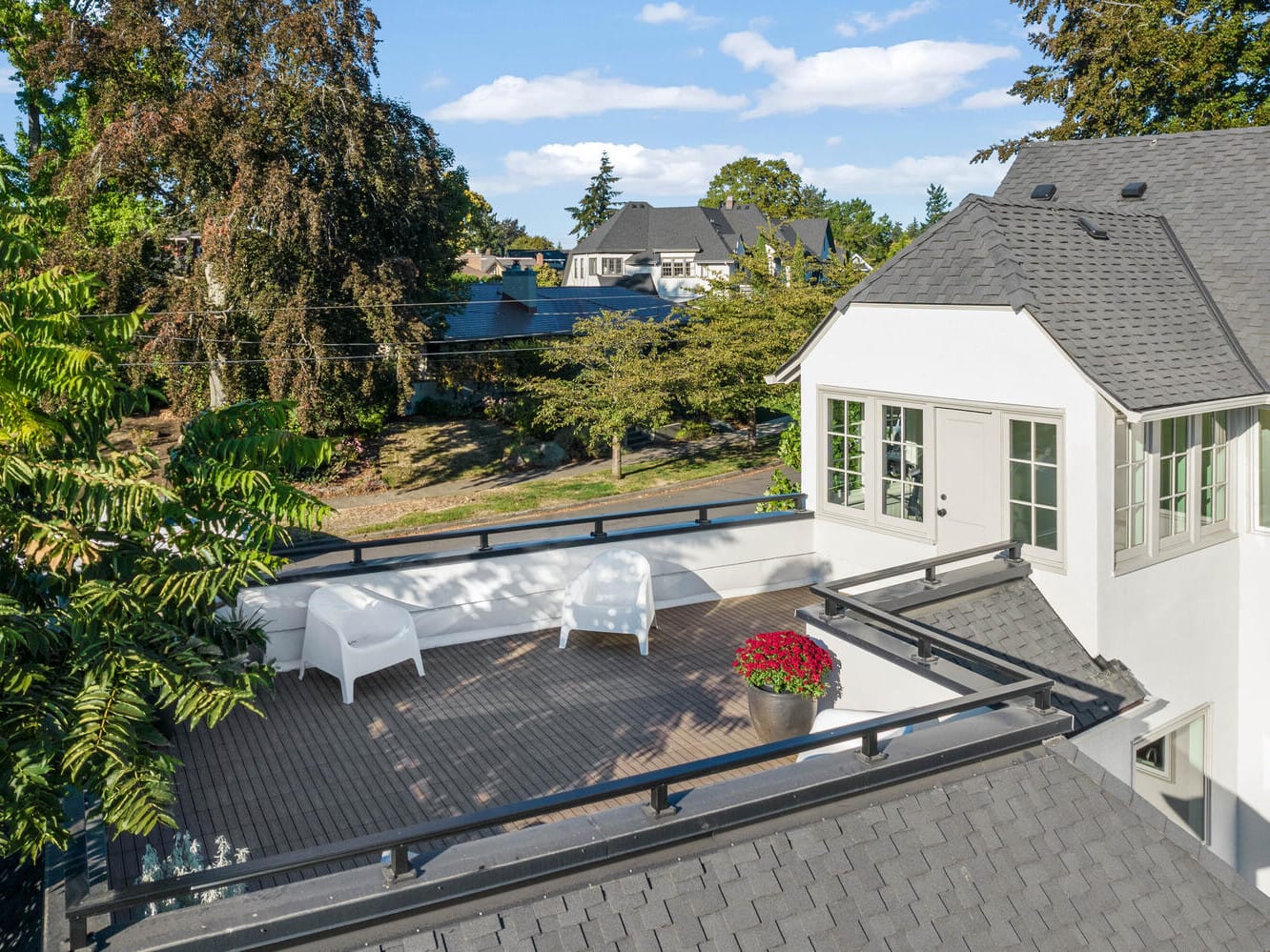 A rooftop patio in Portland, Oregon, features two white chairs and a potted plant with red flowers. Surrounded by trees, it overlooks a charming suburban neighborhood with houses in the background under a partly cloudy sky.