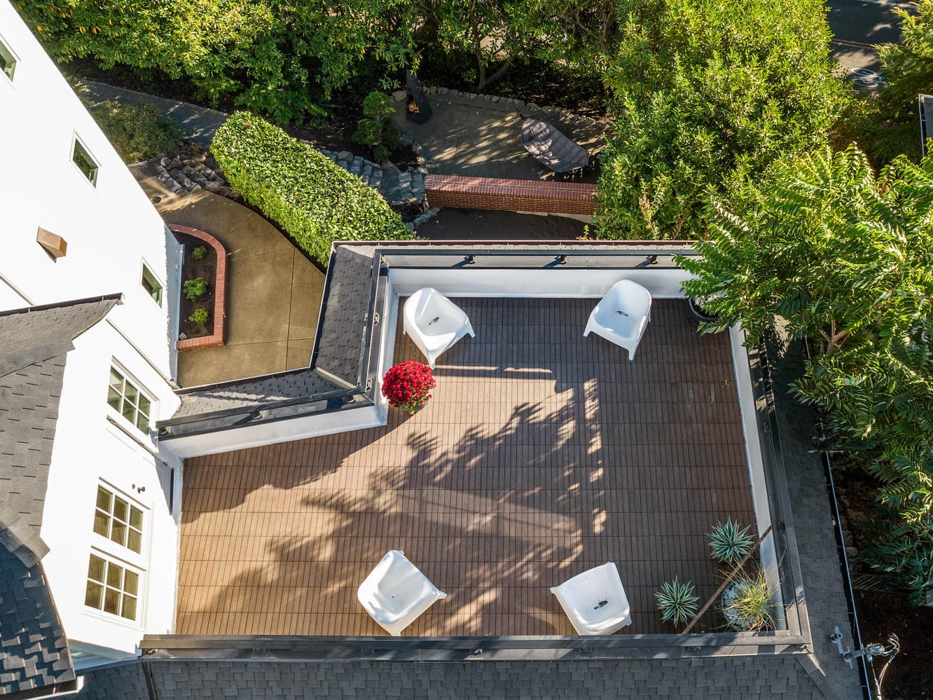 Aerial view of a rooftop patio in Portland, Oregon, featuring four white chairs and a red potted plant on wooden decking. Surrounded by leafy trees and neighboring buildings' roofs, casting shadows on the patio. A narrow path with greenery is visible below.