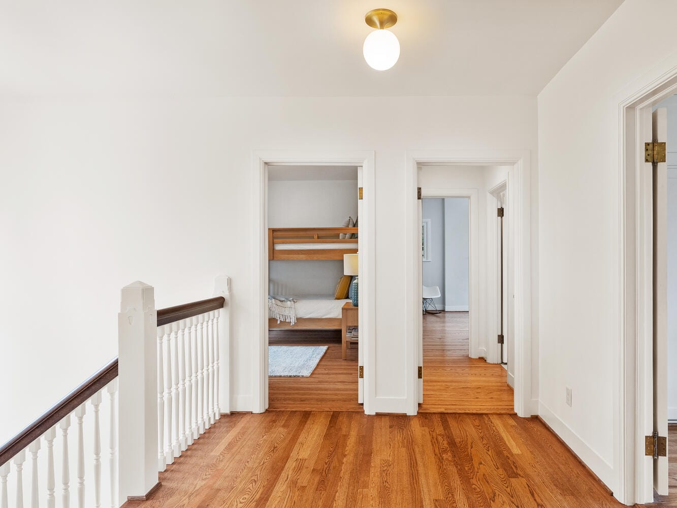 A bright hallway in a Portland, Oregon home features polished wooden floors and white walls. Two open doors lead to rooms, one of which contains a bunk bed. A simple light fixture hangs from the ceiling, and the staircase railing is visible on the left.