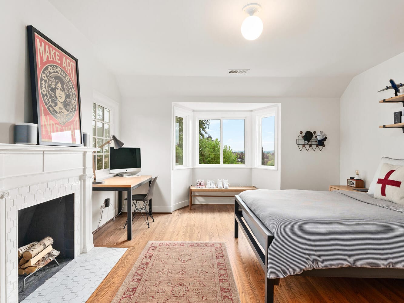 A bright bedroom in a Portland, Oregon style features a bed, framed artwork above a white mantel, and a desk with a computer by the bay window. Shelves display decor while the hardwood floor showcases a patterned rug, giving the room its minimalistic, cozy feel.