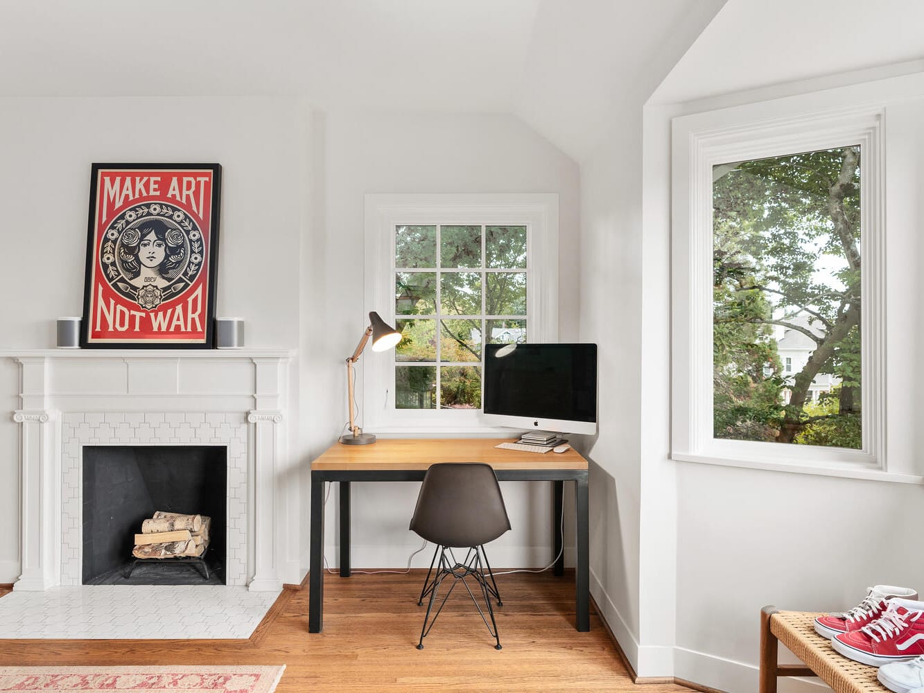 A tidy and bright Portland, Oregon home office features a desk with a computer, lamp, and chair next to a window with a garden view. A white fireplace with logs, modern art saying 