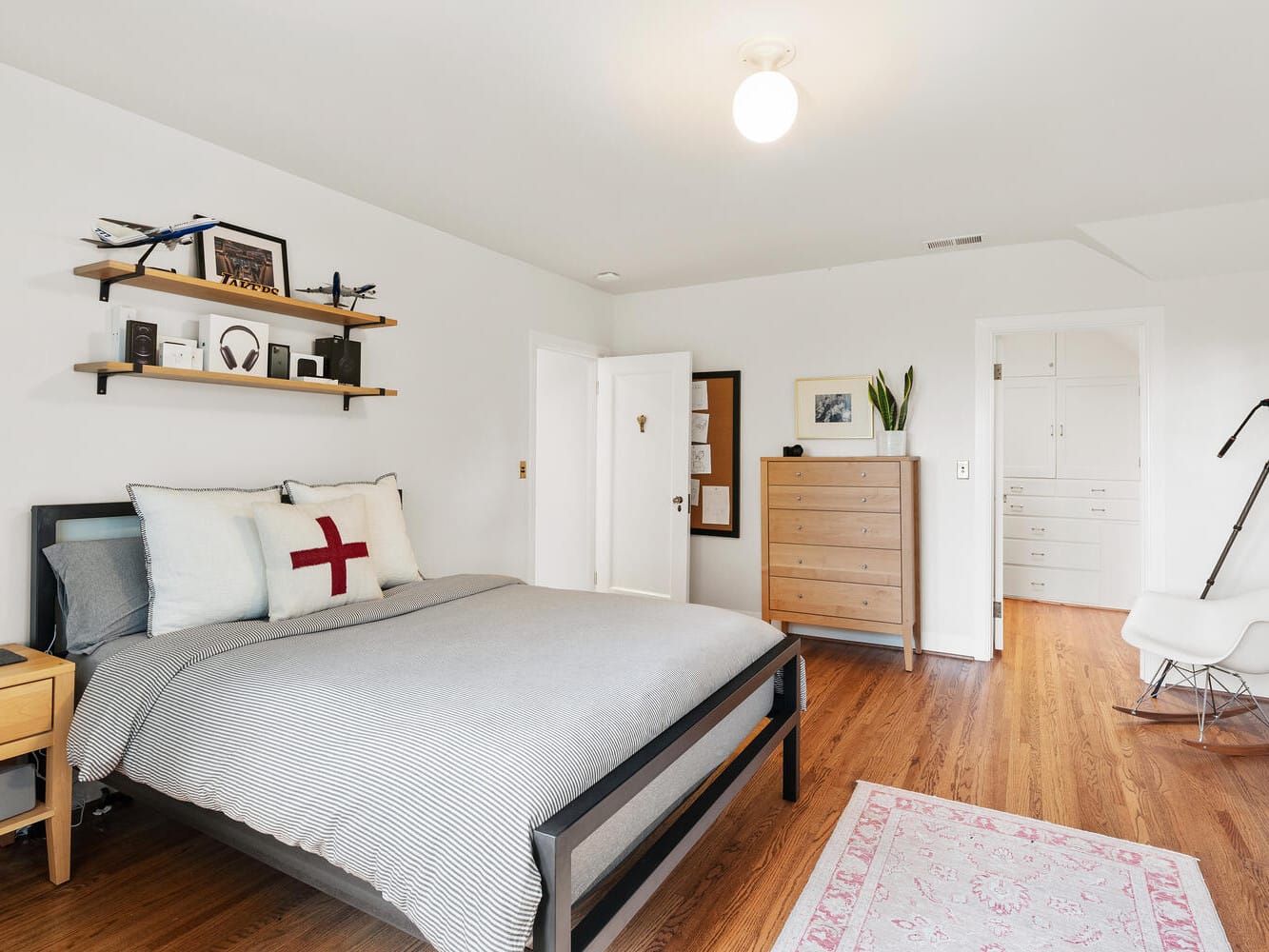 A modern Portland, Oregon bedroom boasts a neatly made bed with a gray comforter. Shelves with decorative items sit above, while hardwood floors accentuate the wooden dresser, tripod, rocking chair, and rug. White walls and a ceiling light complete the serene space.