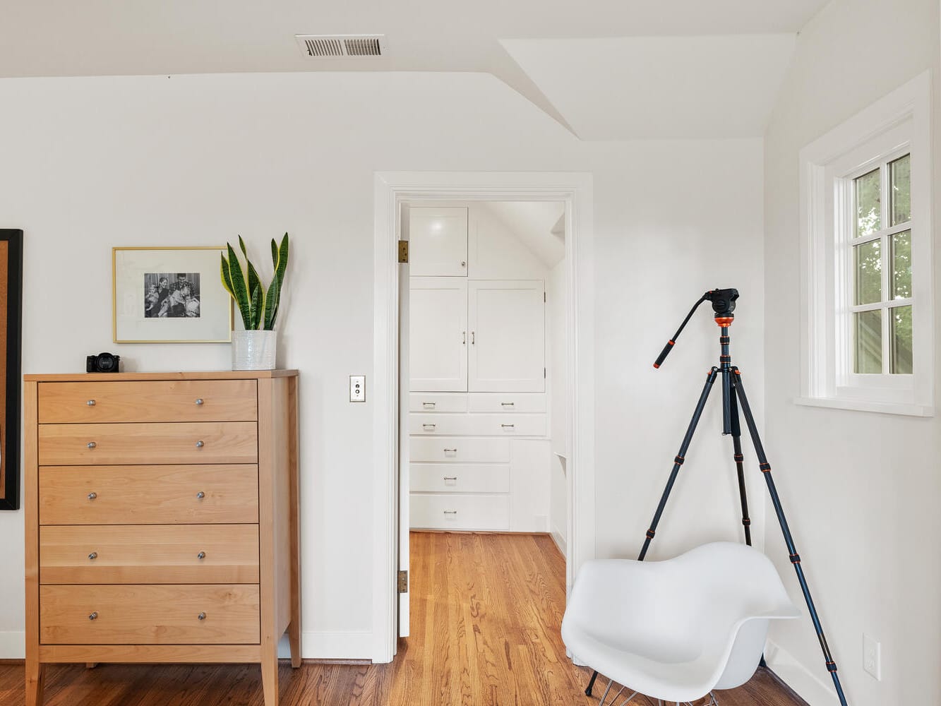 A bright room in Portland, Oregon, features a wooden chest of drawers, a small plant, and pictures on the wall. A camera tripod and a white modern chair are nearby. An open doorway leads to a white closet area with built-in cabinets. Wood flooring throughout.