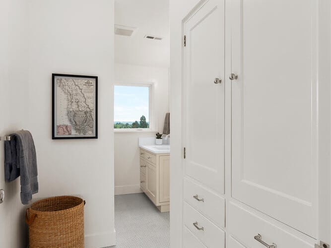 A bright bathroom in Portland, Oregon features white cabinets and drawers, a wicker laundry basket, and a towel hanging on the wall. A framed map hangs above the basket, while a window fills the space with natural light, illuminating hexagonal floor tiles.