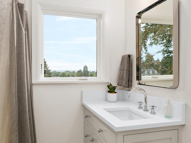 A bright Portland, Oregon bathroom features a white vanity and sink with a mirror above, plus a window framing the lush outdoors. A potted plant adds charm on the counter. The room includes a shower with a curtain and classic small hexagonal tile flooring.