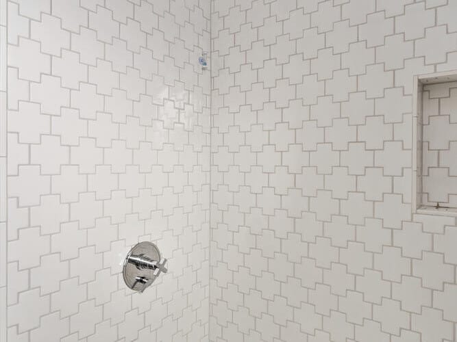 A modern bathroom shower in a Portland, Oregon home features white tiles with a geometric pattern. The stainless steel showerhead, faucet, and handles provide sleek touches, while a small recessed shelf adds convenience. The white tub perfectly complements the clean design.