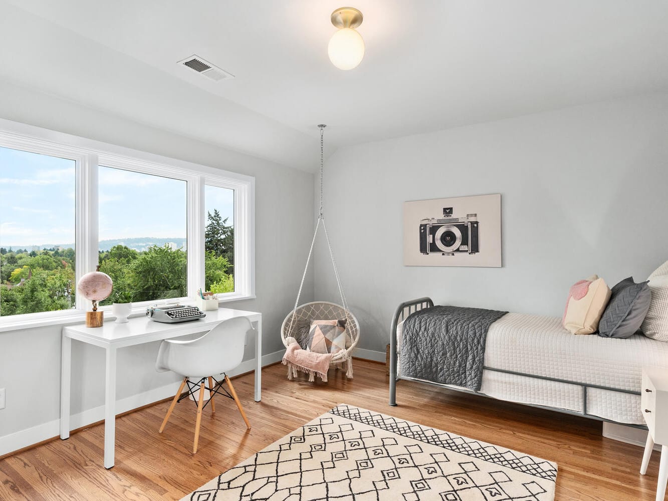 A bright, minimalist bedroom in Portland, Oregon boasts a window view of trees. It features a white desk with a typewriter, a gray bed with a pink pillow, a round hanging chair, and a black-and-white camera photo on the wall. The hardwood floor is adorned with a geometric rug.