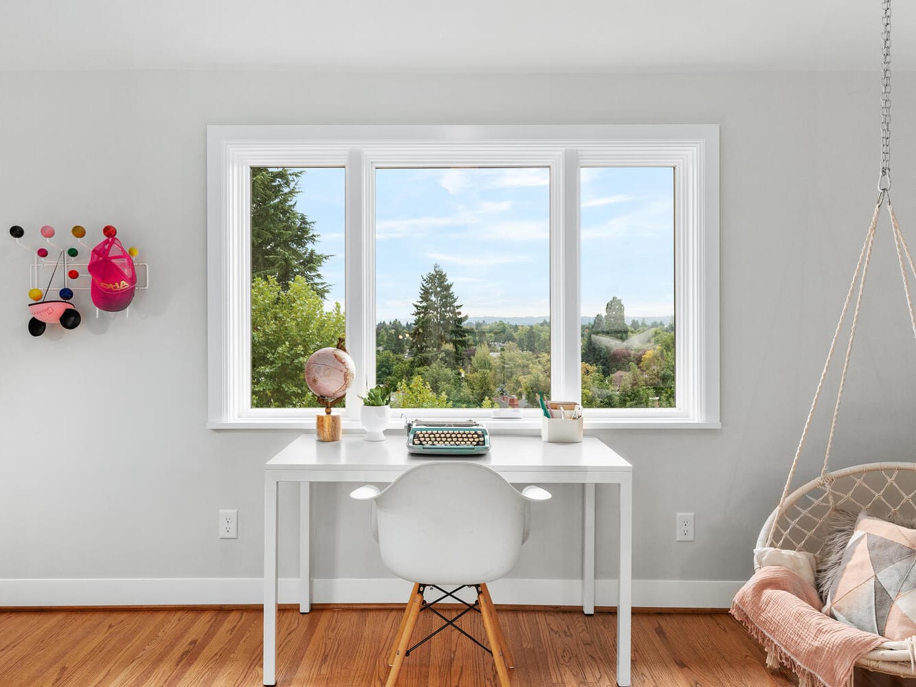 A bright room with a large window view showcasing Portland, Oregon's lush trees and blue sky. Inside, there’s a white desk with a pink globe and typewriter, a white chair, colorful wall art, and a hanging chair adorned with a pink pillow and blanket.