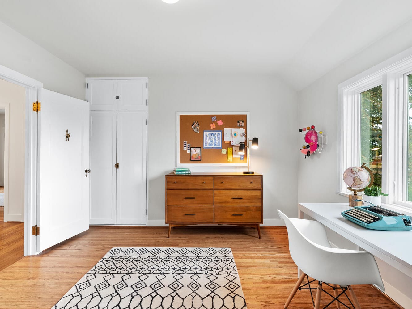 In a bright Portland, Oregon room with wooden flooring, a white desk and chair accompany a typewriter by the window. A patterned rug adds flair beneath. Against the wall sits a wooden dresser with a corkboard above, while to the left, a closed white door stands quietly.