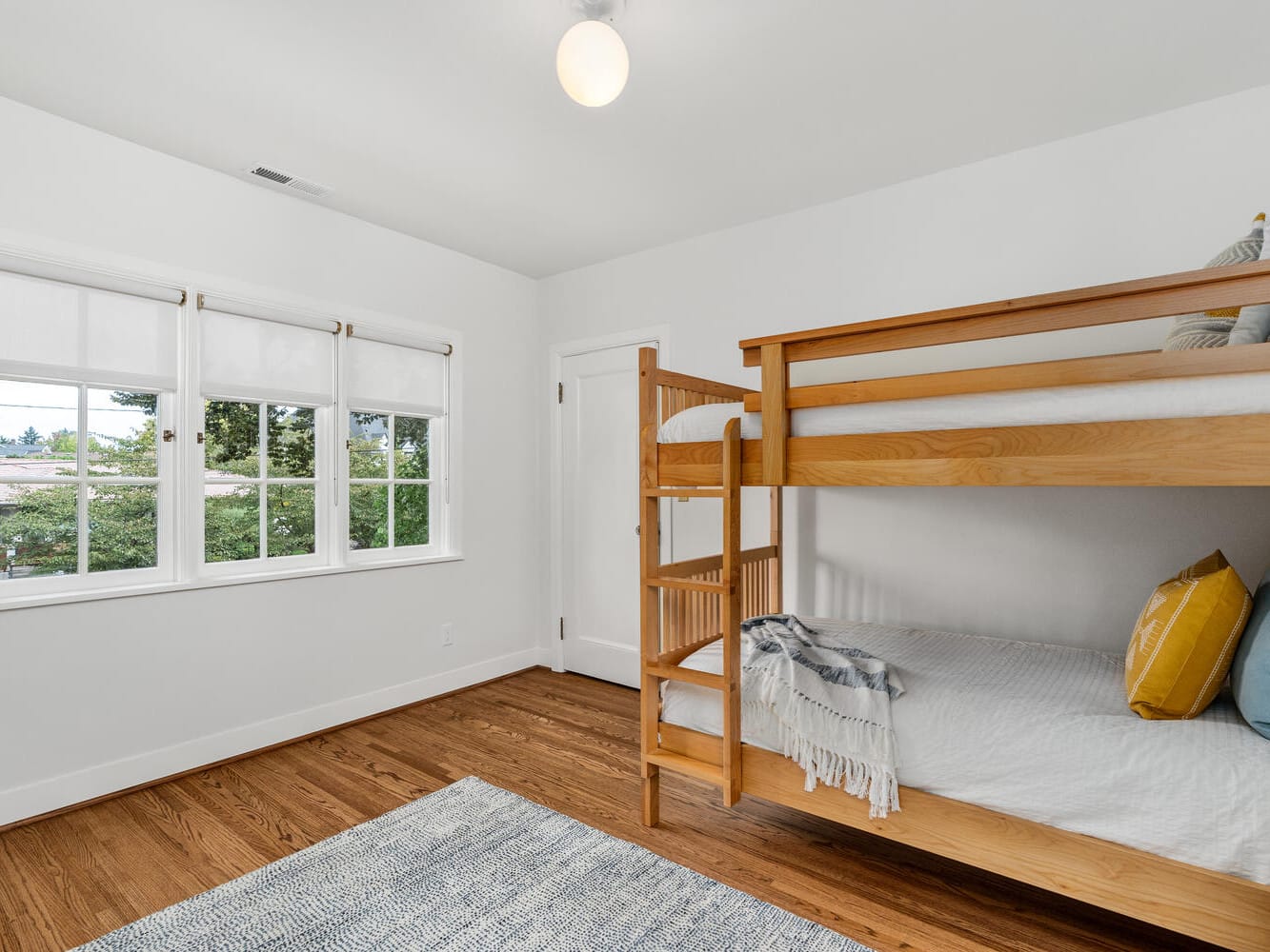 A clean, minimalist bedroom in Portland, Oregon features wooden bunk beds with white and blue bedding. Natural light streams in through large windows with white blinds. The room has wooden flooring and a small gray rug, creating a cozy, inviting atmosphere.