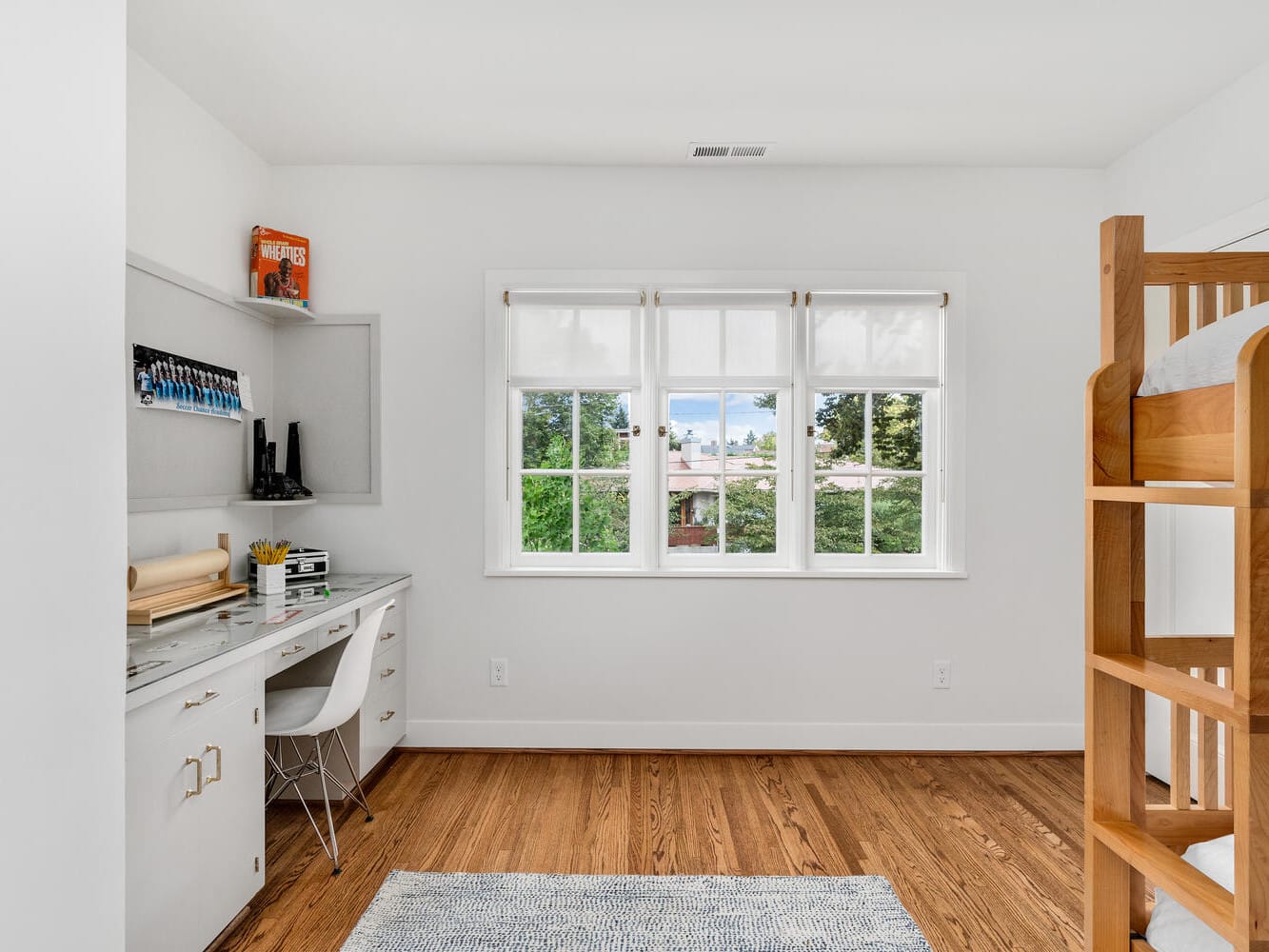 A bright, minimalist room in Portland, Oregon features a wooden bunk bed on the right and a small desk with a chair on the left. The space boasts hardwood floors, a large window with views of trees, and a few decorative items adorning the desk and shelf.