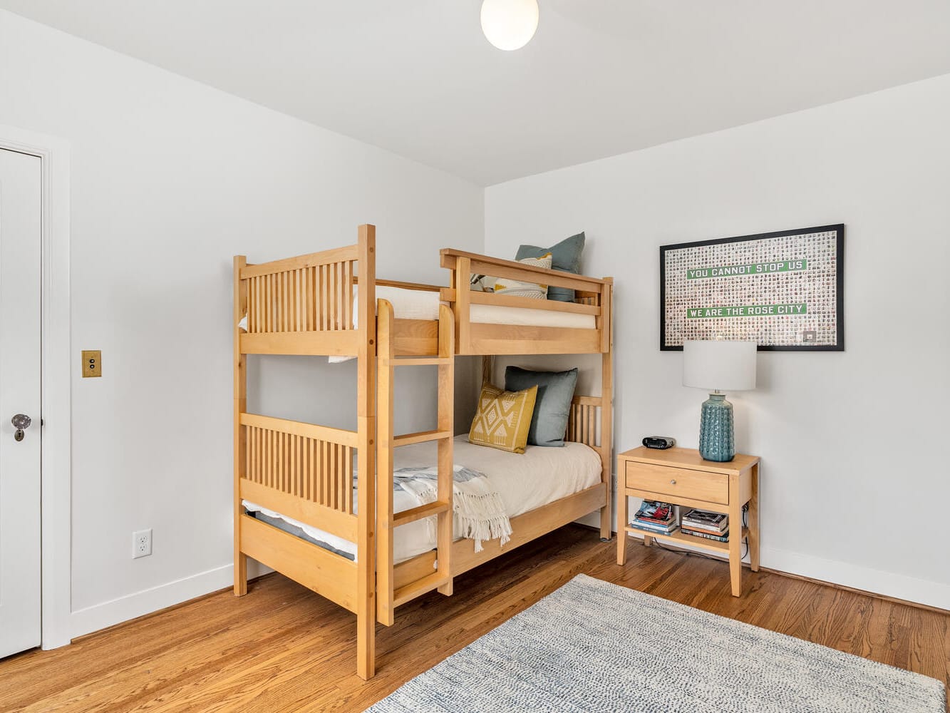 A cozy Portland, Oregon, bedroom featuring wooden bunk beds with white and pastel bedding, a small wooden nightstand with a lamp and decor, and a framed art piece on the wall. The room has hardwood floors and a soft gray rug.